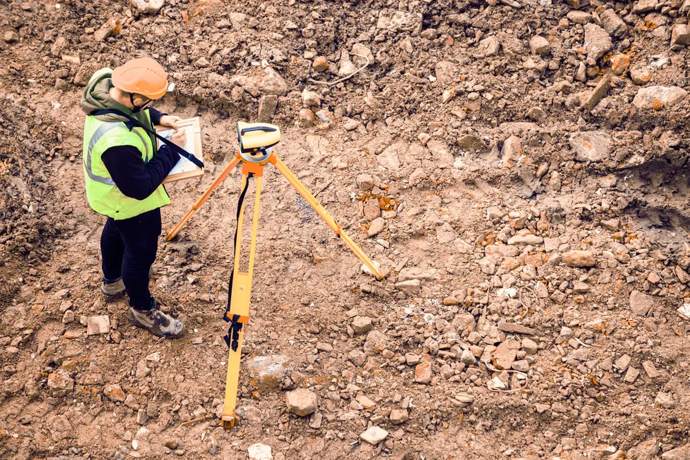 A surveyor in a safety vest and hard hat with a tripod on rocky terrain