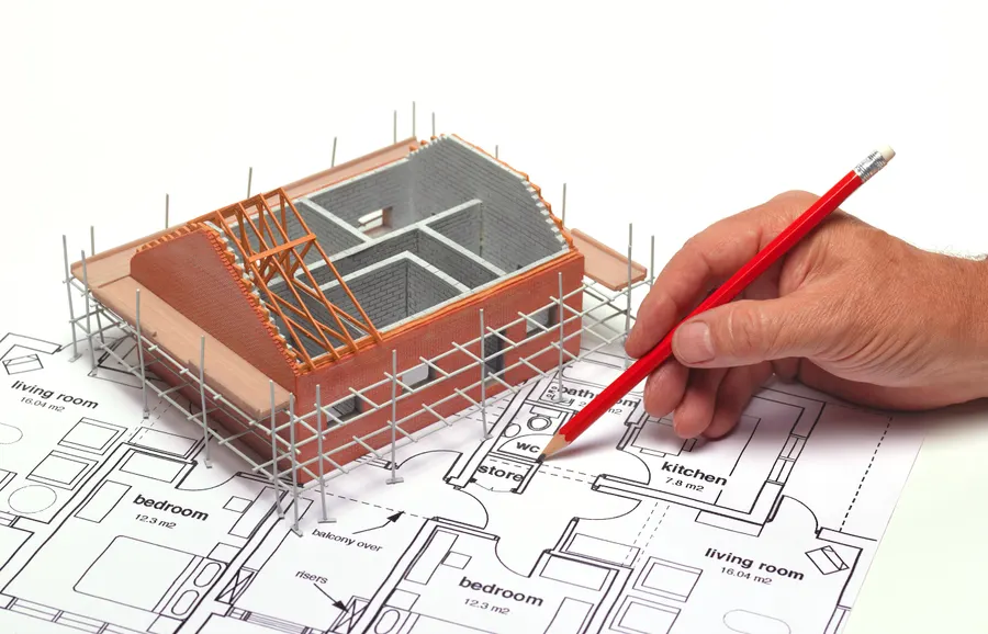 A hand holds a red pencil over architectural plans beside a 3D model